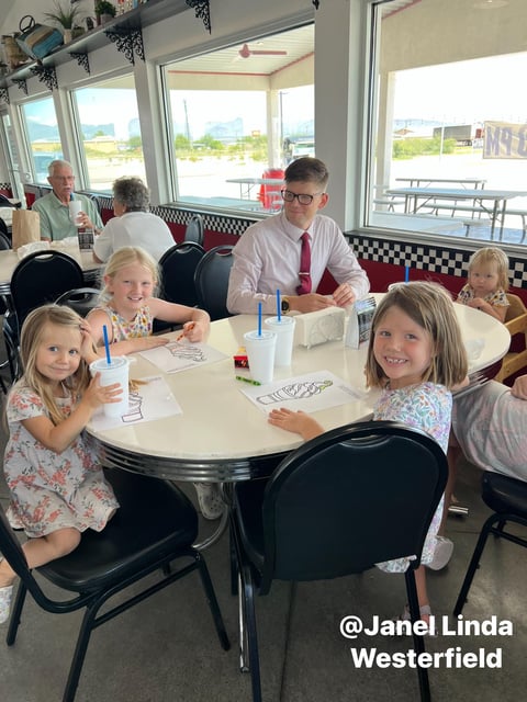 Family dining at a retro diner with children seated at a round table, an adult standing nearby, and large windows showing a waterfront view.