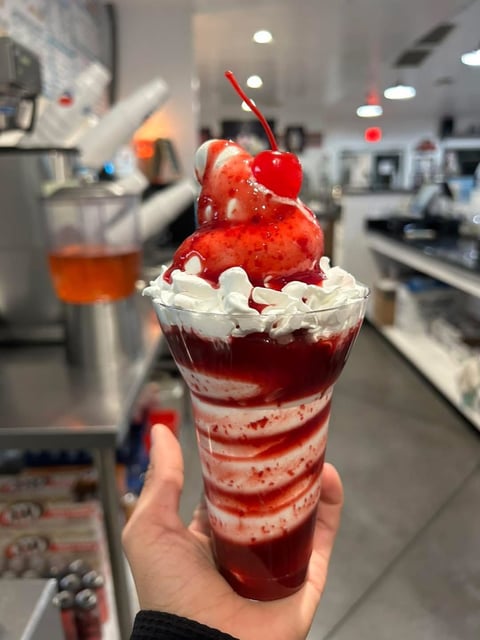 Hand holding a layered red and white sundae with whipped cream, strawberry, and cherry on top in an ice cream shop