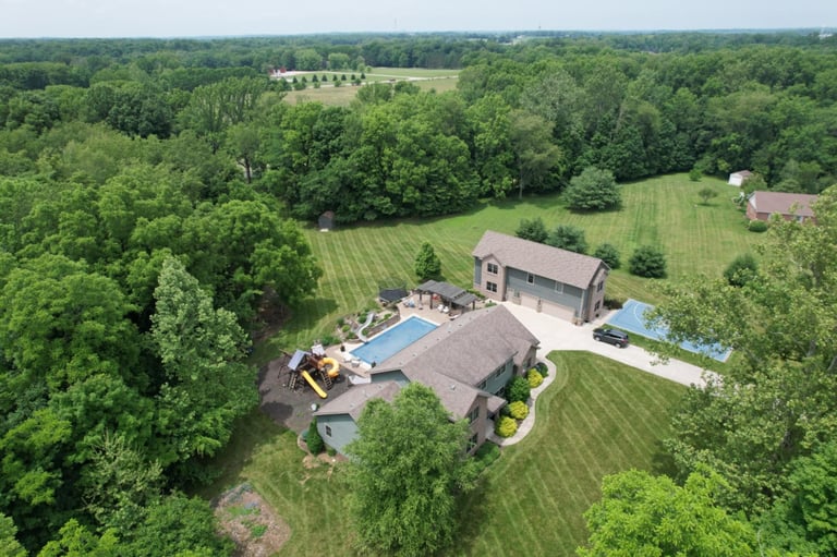 Aerial view of a residential home with swimming pool surrounded by lush green lawns and wooded landscape