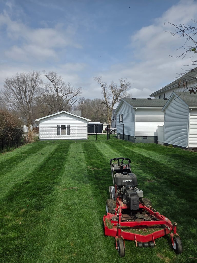 Red zero-turn mower on striped lawn with white cottage and houses in background under blue sky