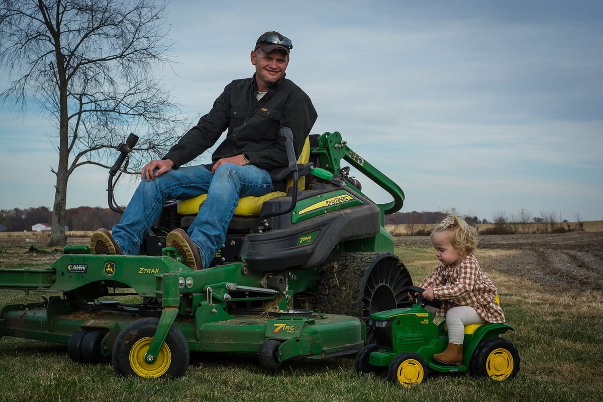 Man sitting on green and yellow riding lawn mower with child on toy tractor in rural field
