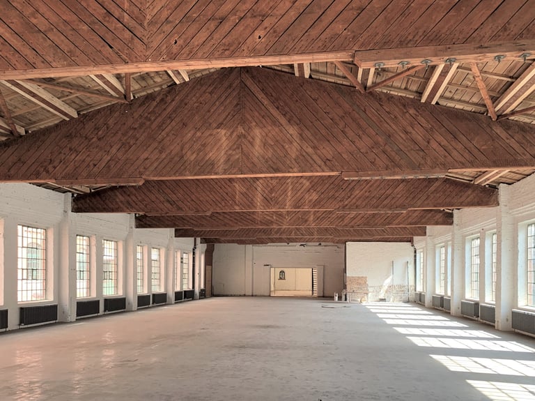 Leerstehende Hallenfläche mit freiliegender Holzbalkendecke, weißen Wänden und großen Fenstern entlang beider Seiten