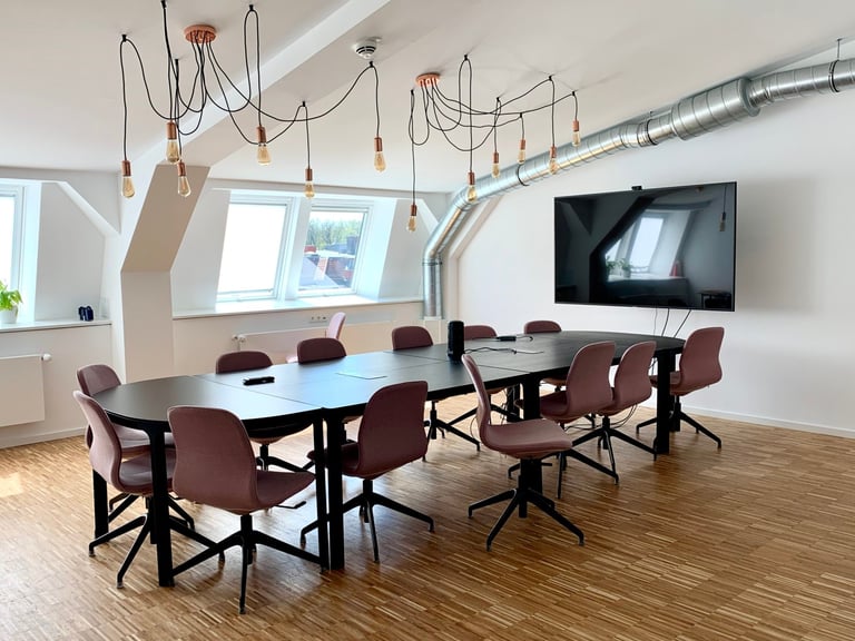 Moderner Konferenzraum mit langem Tisch, burgunderroten Stühlen, wandmontiertem Bildschirm, dekorativen Pendelleuchten und hellen Fenstern
