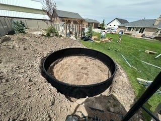 Large black ring-shaped tank filled with sand in a residential backyard with buildings and landscaping visible in the background