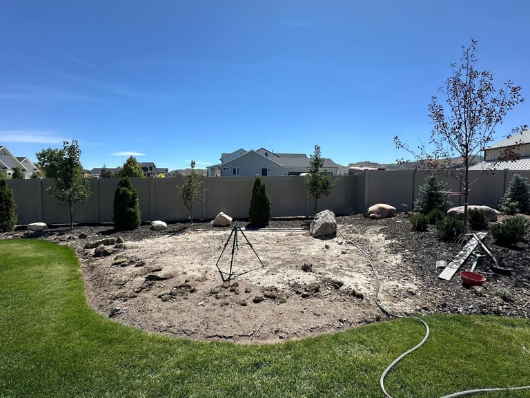 Backyard landscape under construction with cleared dirt area, surveying tripod, scattered rocks, young trees, green grass strip, and gray fence under blue sky