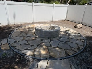 Circular stone fire pit with seating wall in center, surrounded by flagstone patio with white fence in background