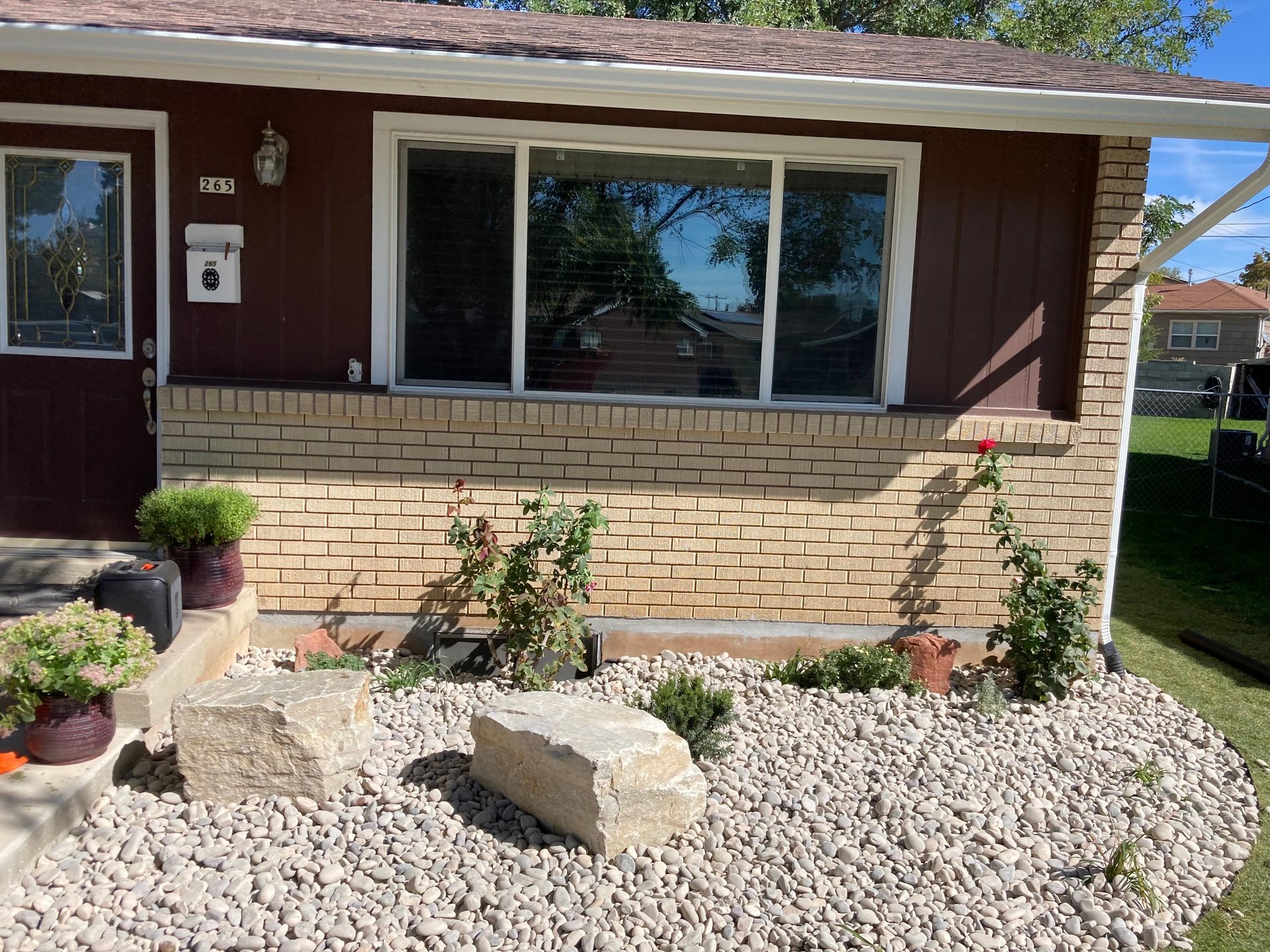 Front of brick house with large windows, gravel landscaping, potted plants, and climbing vines in sunny yard