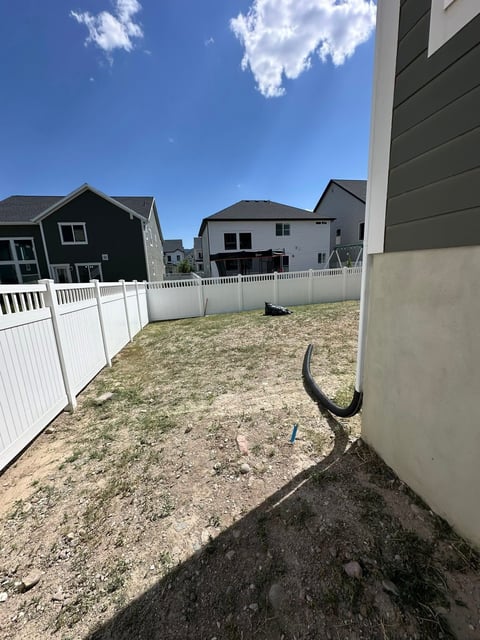 Residential backyard with white fence, sparse grass, and neighboring houses under blue sky