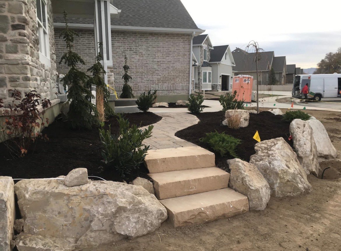 Landscaped front yard with stone steps, large boulders, and shrubs beside brick house