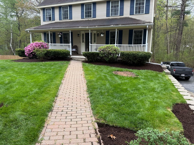 Two-story colonial house with brick walkway, manicured lawn, white railings, and pink flowering shrubs