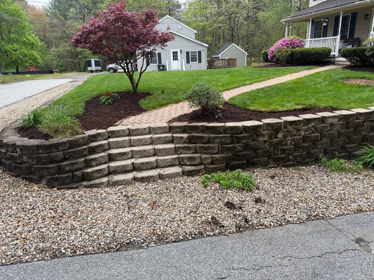 Residential front yard with stacked stone retaining wall, gravel landscaping, and magenta flowering tree near white house