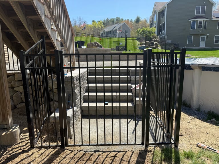 Black metal gate with vertical bars open between two fenced sections of a residential yard with houses visible in the background