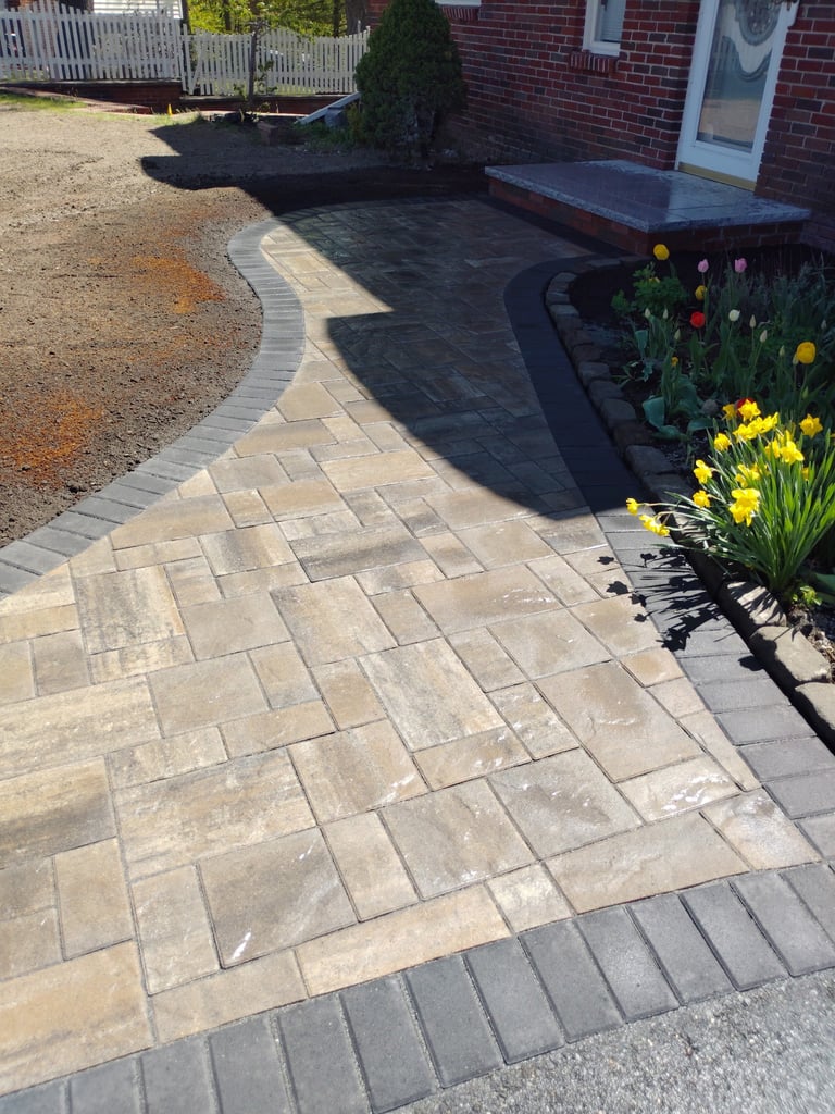 Residential driveway with beige paver entrance, black asphalt section, brick home, and colorful flower garden with yellow and red blooms