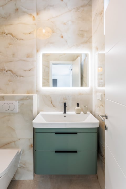 Modern bathroom with illuminated mirror, cream marble walls, white vessel sink, and green vanity cabinet with two drawers