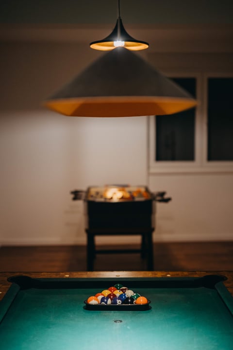 Billiard table with balls racked and a pendant lamp illuminating the green pool table surface in an indoor game room