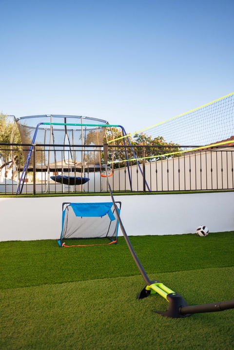 Backyard play area with trampoline, soccer goal, volleyball net, and ball on artificial grass