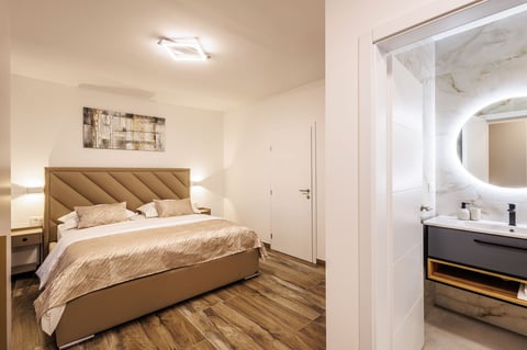 Modern bedroom with tan upholstered bed, wooden nightstands, and an open doorway to a contemporary bathroom with illuminated mirror