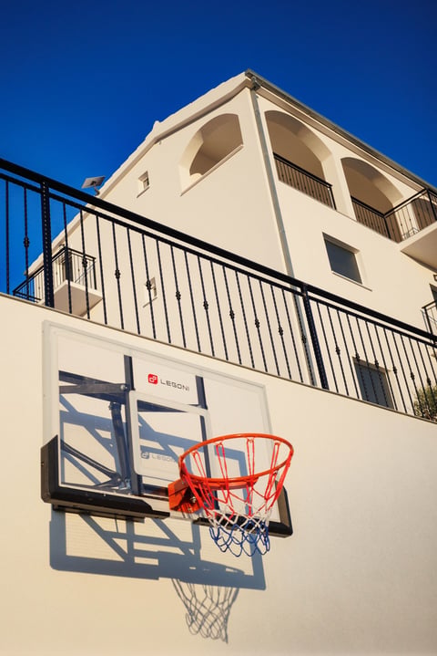 Basketball hoop mounted on cream-colored building wall with metal railing and balcony above, blue sky background