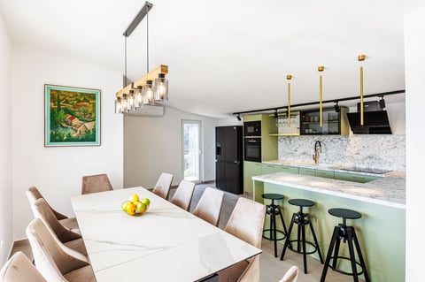 Modern kitchen and dining area with white table, tan chairs, green kitchen cabinets, black bar stools, and pendant lighting