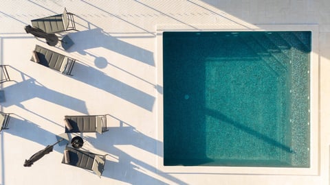 Overhead view of lounge chairs and an umbrella casting shadows on a white deck beside a turquoise swimming pool