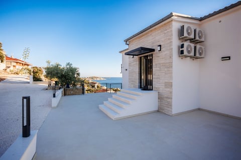 Modern white stone villa with terrace overlooking Mediterranean sea, featuring dark entrance door and air conditioning units on facade
