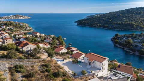 Aerial view of Mediterranean coastal village with whitewashed buildings overlooking turquoise blue water and forested hills