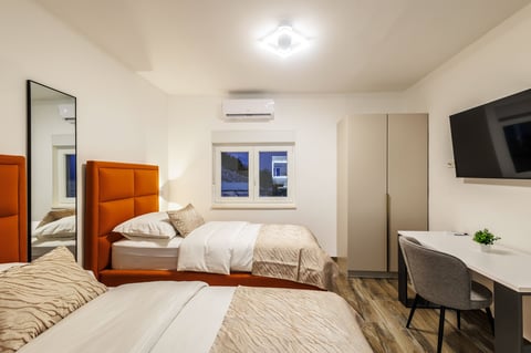 Modern hotel room with two beds, orange headboard, desk, wall-mounted TV, and minimalist furnishings