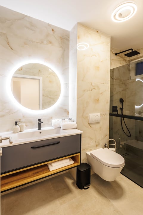 Modern bathroom with illuminated round mirror, floating vanity with wooden shelf, white sink, and glass shower enclosure
