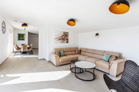 Modern living room with beige L-shaped sectional sofa, round coffee table, pendant lights, and open-concept layout with seating area visible in background