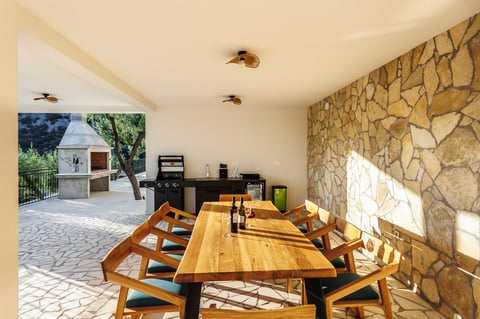 Modern outdoor dining area with wooden table, stone accent wall, and outdoor kitchen with barbecue