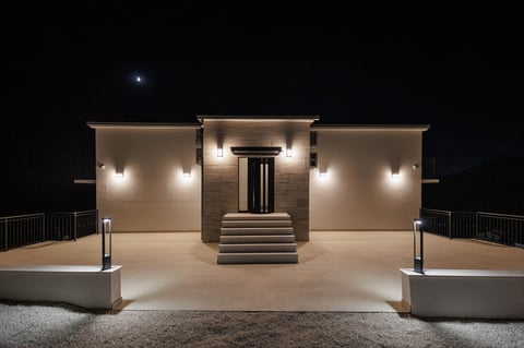 Modern building entrance at night with warm lighting, symmetrical design, central steps, and concrete planters flanking the illuminated facade