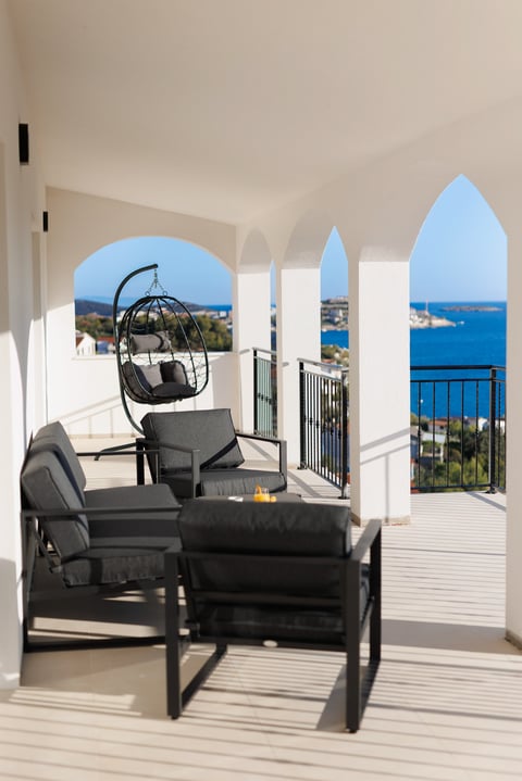 Modern coastal terrace with black lounge chairs and hanging pod chair overlooking blue Mediterranean sea and distant coastline through arched windows