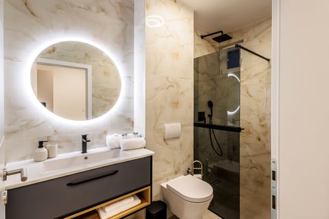 Modern bathroom with illuminated round mirror, marble walls, white sink, dark vanity, and walk-in shower with glass enclosure