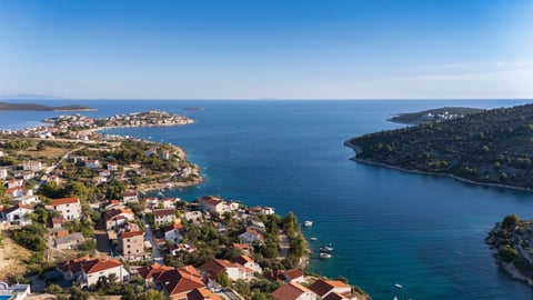 Aerial view of a Mediterranean coastal town with terracotta-roofed houses, turquoise water, and forested hills under clear blue sky