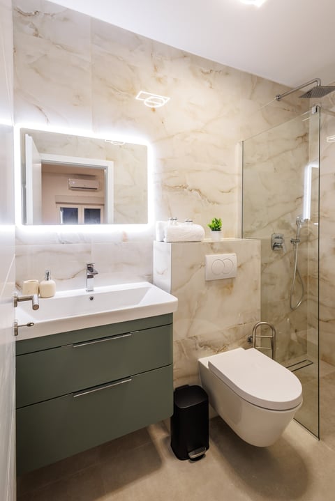 Modern bathroom with marble walls, green vanity, illuminated mirror, glass shower enclosure, and wall-mounted toilet
