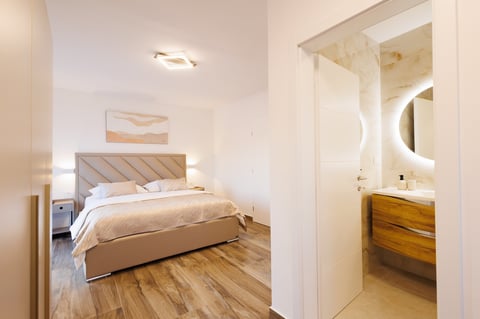 Modern bedroom with wood bed frame, white linens, hardwood floors, and en-suite bathroom visible through open doorway