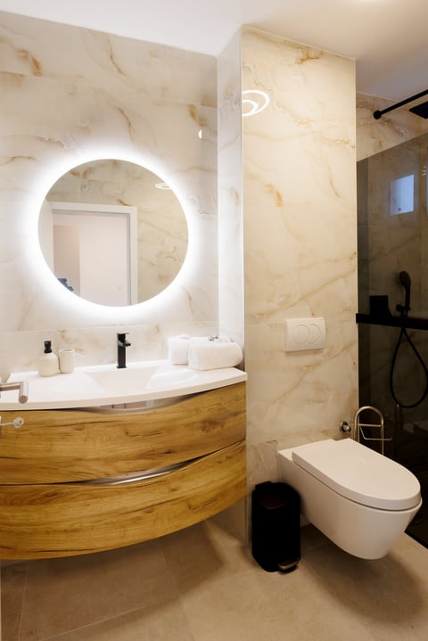 Modern bathroom with marble walls, illuminated circular mirror, wooden vanity with white sink, and wall-mounted toilet with black fixtures