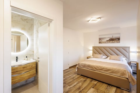 Modern bedroom with beige bed, wooden floor, and en-suite bathroom with illuminated round mirror visible through open doorway