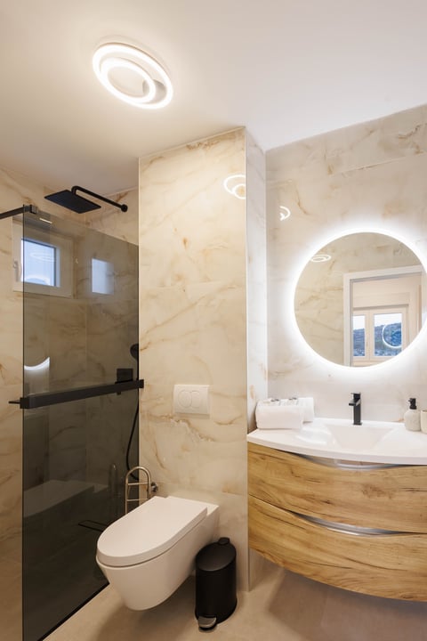 Modern bathroom with marble walls, illuminated circular mirror, wooden vanity, walk-in shower with glass door