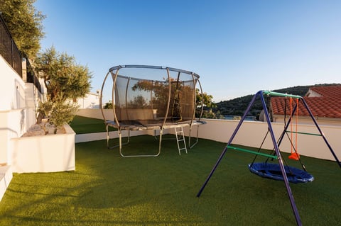 Backyard with round trampoline and blue swing set on artificial grass under clear blue sky