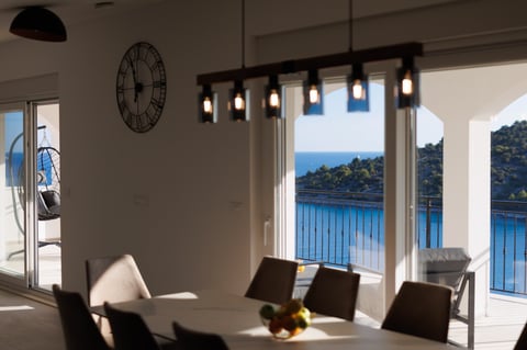 Modern dining room with floor-to-ceiling windows overlooking blue ocean and mountains, featuring black chairs, industrial pendant lights, and a wall clock