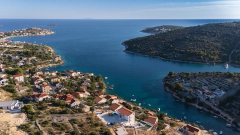 Aerial view of a Mediterranean coastal town with turquoise waters, forested hills, and residential buildings along rocky coves