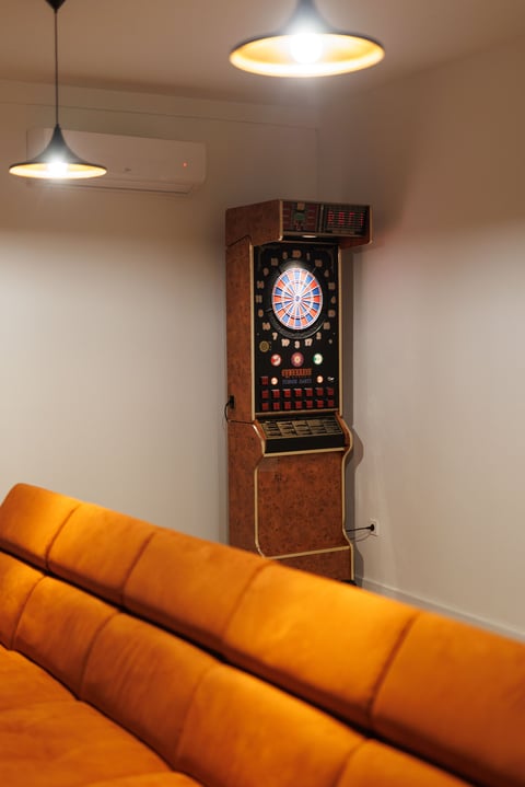 Vintage casino roulette machine in wooden cabinet against white wall, with orange sofa in foreground and pendant lights overhead