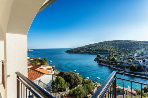 Balcony view of turquoise Mediterranean coastline with white villas, green hillside, and clear blue sky