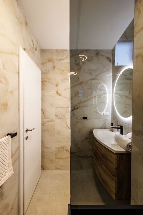 Modern bathroom with marble walls, illuminated round mirror, wooden vanity sink, and white door