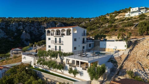 White Mediterranean villa on hillside with terraced courtyard and landscaping, surrounded by rocky terrain and green hillside