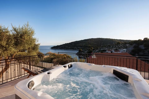Hot tub on wooden deck overlooking a bay with forested hills and blue water under clear sky
