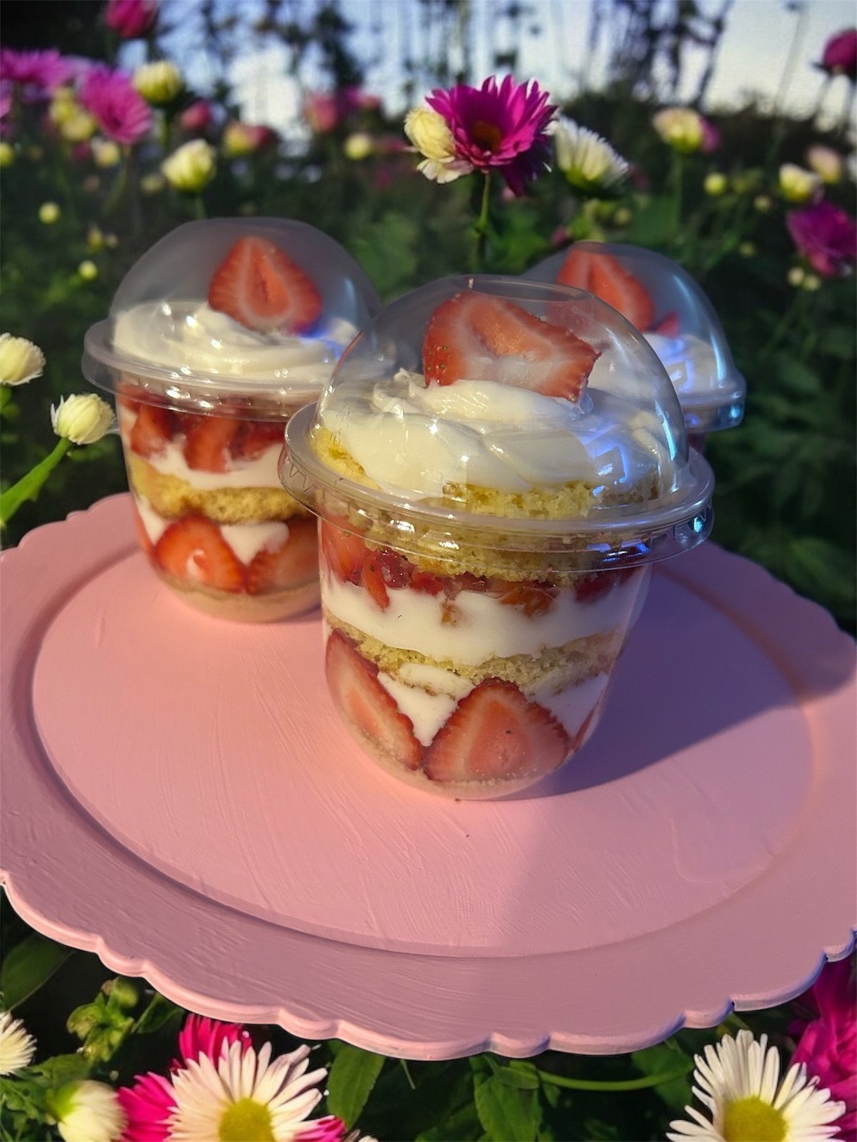 Three layered strawberry shortcake desserts in clear cups on a pink plate surrounded by colorful daisies in a flower garden