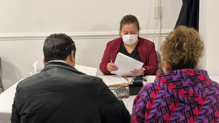 Woman wearing a face mask and burgundy cardigan holds papers while seated across from two other people at a table