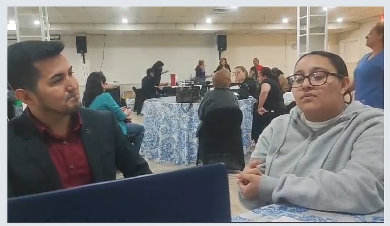 Two men having a conversation at an indoor event or gathering with other people and tables visible in the background